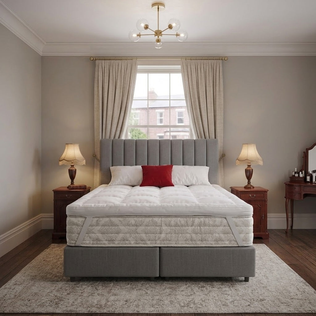 Grey bed with white mattress topper strapped to the off white mattress, red pillow, and wooden lamps on both sides of the bed in a bedroom setting.