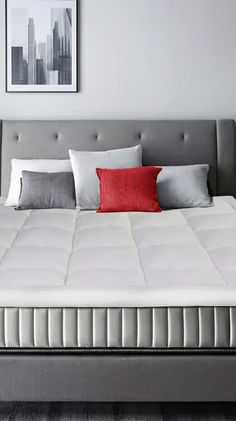 Gray bed with a white mattress topper on top with gray and red pillows against a white wall.