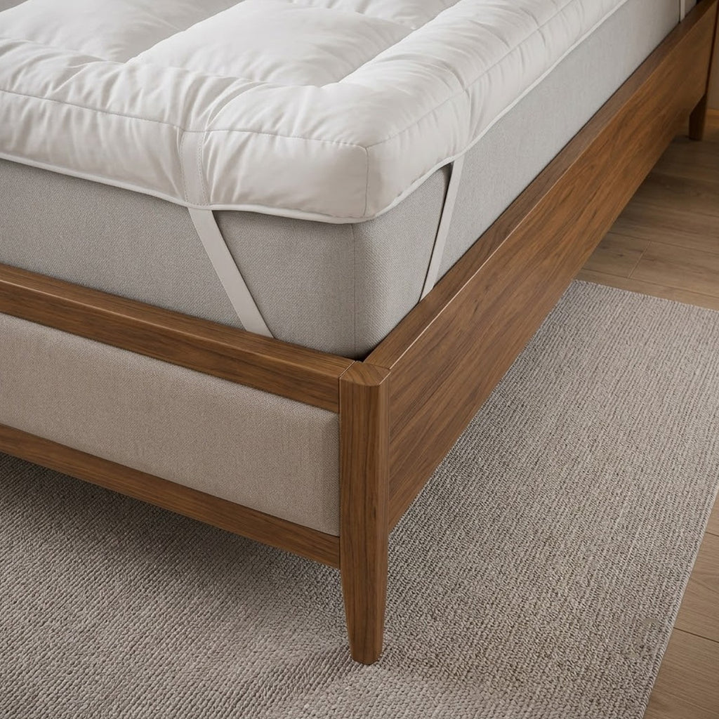 A close-up of a bed frame with a white mattress topper, shown in a styled bedroom with wood flooring and a rug.